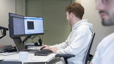 Au premier plan, un homme est assis à son bureau, partiellement cadré et vu de profil, le regard fixé droit devant lui, comme s’il observait un écran d’ordinateur. À l’arrière-plan, un autre homme, vu de profil, tient une souris à la main devant deux ordinateurs ; il regarde attentivement celui de droite.