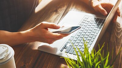 Une personne qui tient son téléphone dans une main et touche son sur l'ordinateur portable avec son autre main