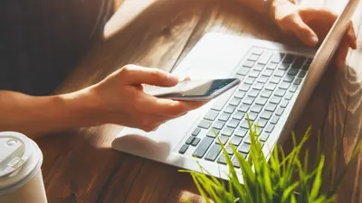 Une personne qui tient son téléphone dans une main et touche son sur l'ordinateur portable avec son autre main