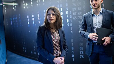 two professionals, a woman and a man, in a high-tech environment with digital data or code displayed on the wall behind them, suggesting a setting related to cybersecurity, data analysis, or technology.