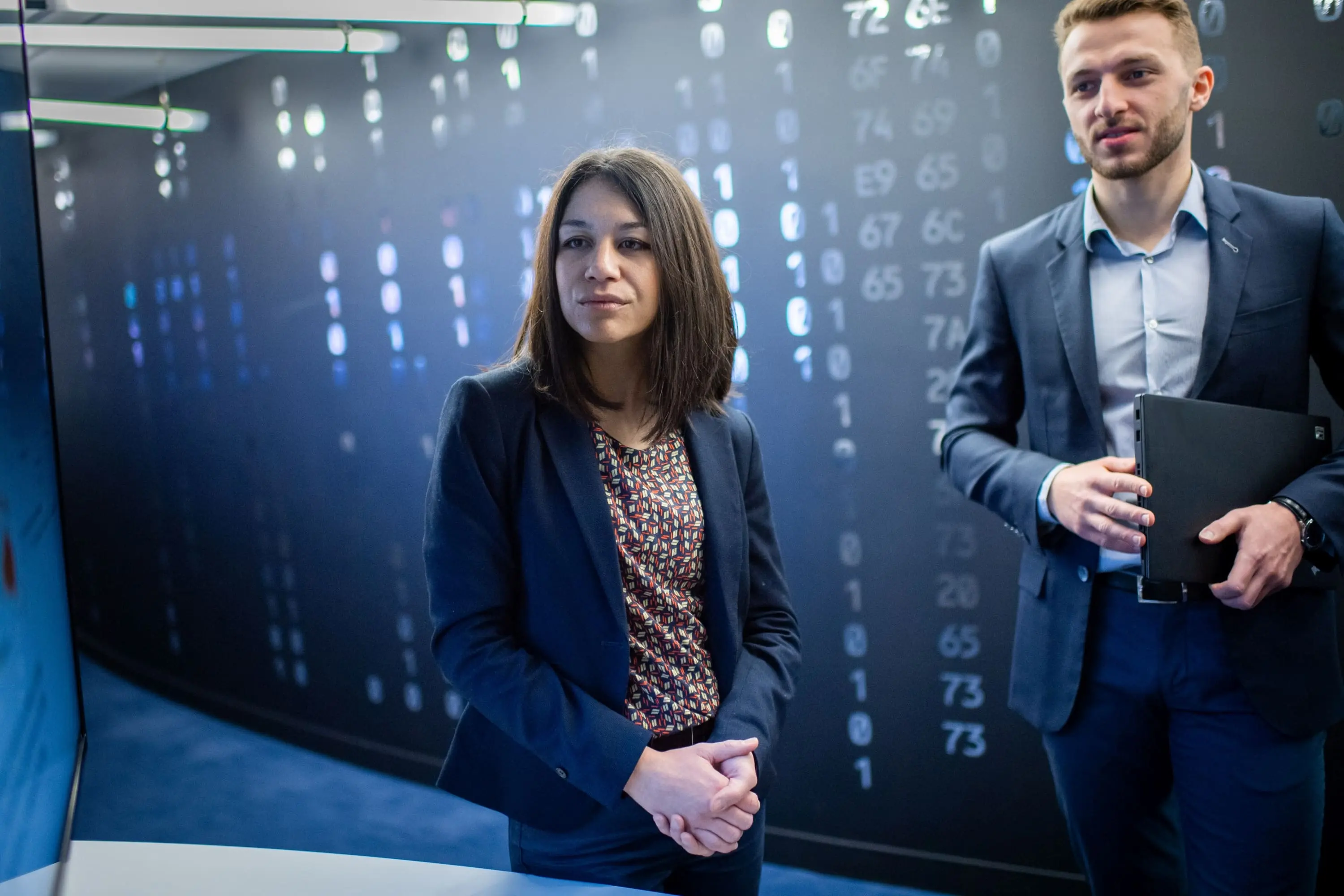 two professionals, a woman and a man, in a high-tech environment with digital data or code displayed on the wall behind them, suggesting a setting related to cybersecurity, data analysis, or technology.