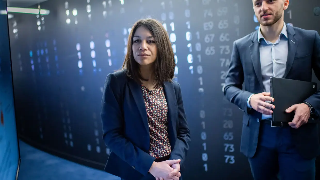 two professionals, a woman and a man, in a high-tech environment with digital data or code displayed on the wall behind them, suggesting a setting related to cybersecurity, data analysis, or technology.