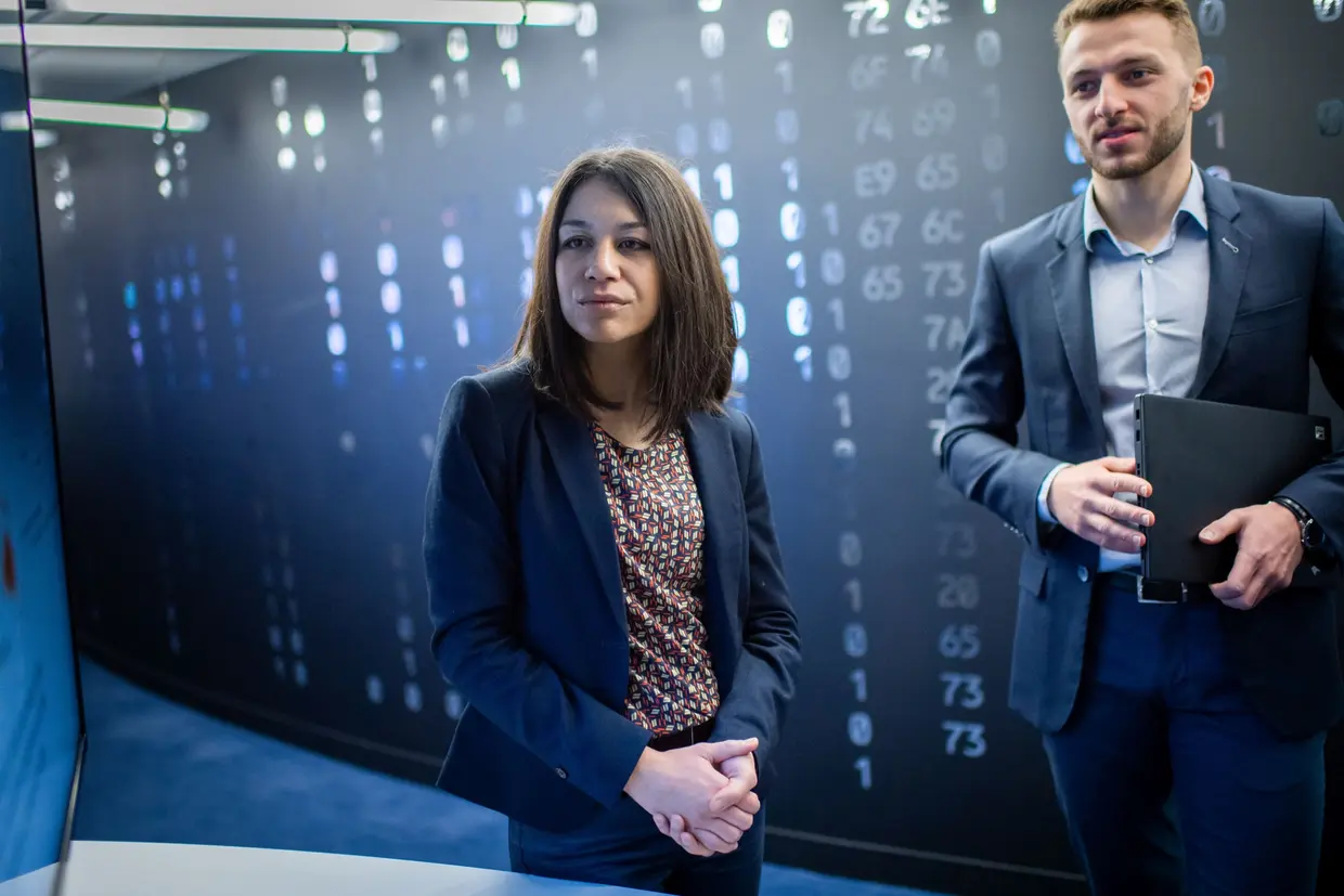 two professionals, a woman and a man, in a high-tech environment with digital data or code displayed on the wall behind them, suggesting a setting related to cybersecurity, data analysis, or technology.