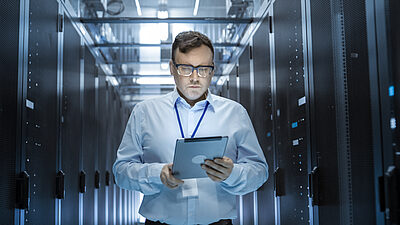 Un homme portant des lunettes, une chemise et une tablette marchant au milieu d'une salle de serveurs informatique. 