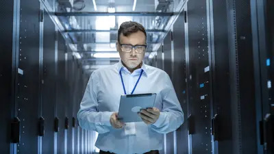 Un homme portant des lunettes, une chemise et une tablette marchant au milieu d'une salle de serveurs informatique. 