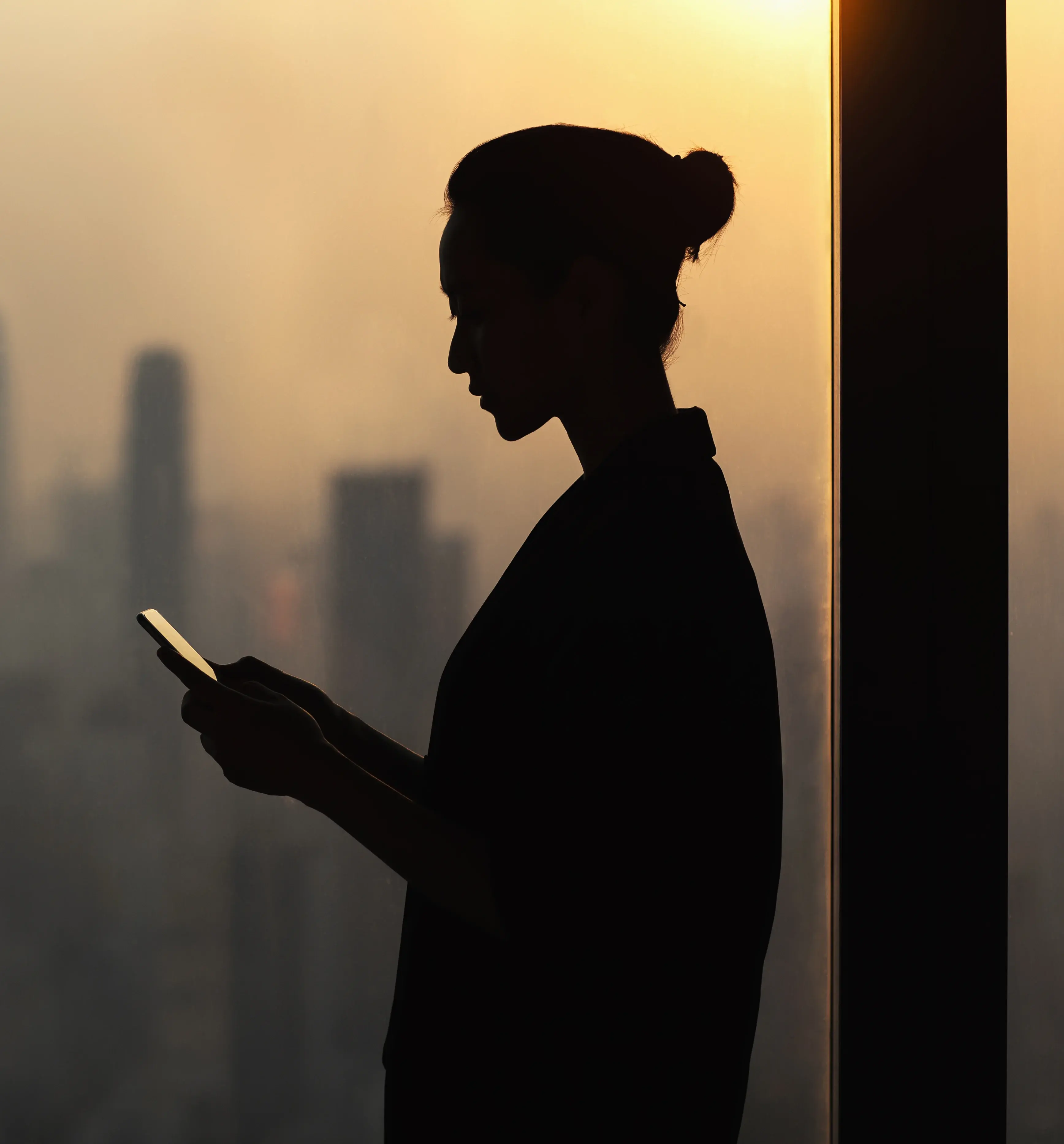 Silhouette of a woman, next to a window, looking at a tablet with a city skyline in the background