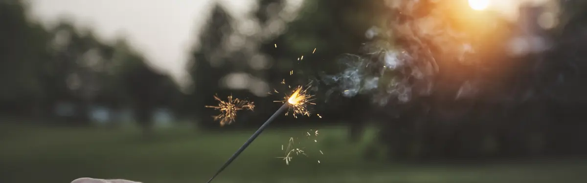 Hand holding a sparkler, lit and glowing, outdoors at sunset or dusk.