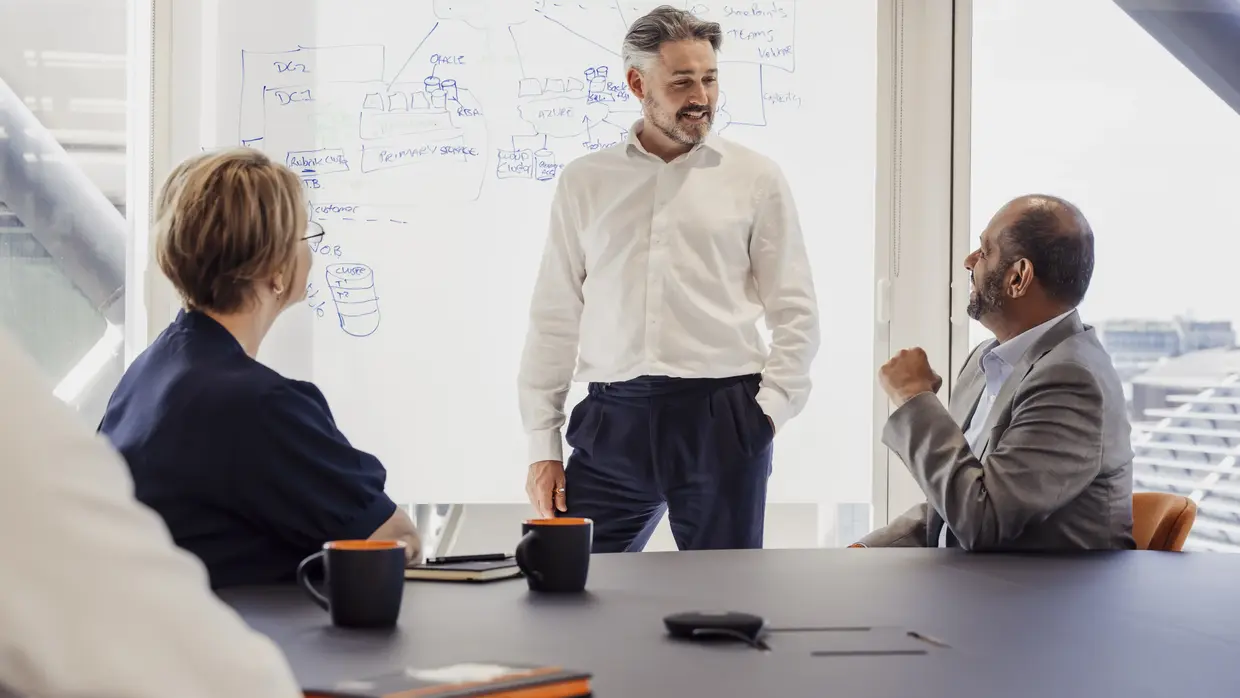 „Ein Mann steht und spricht vor einer Gruppe von drei Personen in einem modernen Besprechungsraum mit großen Fenstern und einer Whiteboard-Wand im Hintergrund.