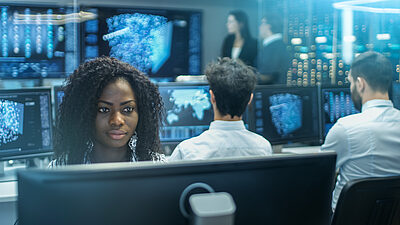 A woman with other team members in the background, in a control center with data models on the screens