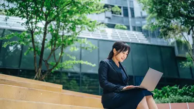 Eine Frau arbeitet im Freien an einem Laptop. Im Hintergrund sind Bäume und ein modernes Gebäude zu sehen.