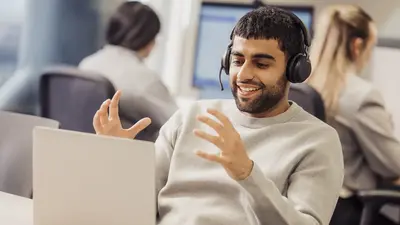 Ein Mann mit Headset sitzt an einem Tisch, lächelt und gestikuliert beim Gespräch. Im Hintergrund sind zwei weitere Personen an Computern, die sich auf ihre Bildschirme konzentrieren.