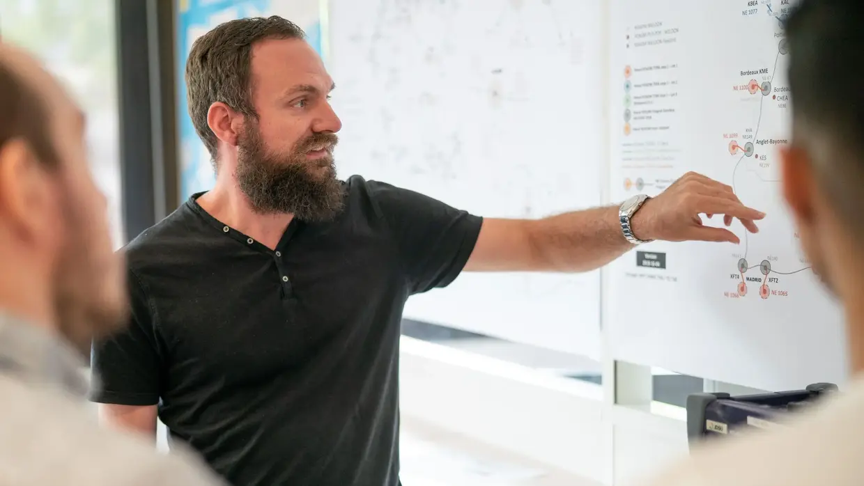 A  man giving a presentation to a group, pointing at a large diagram on a whiteboard, in a professional setting.