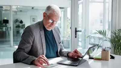 Älterer Mann mit weißen Haaren und Brille sitzt an einem Schreibtisch in einem modernen Büro. Er trägt einen grauen Anzug und eine dunkle Shirt. Er arbeitet mit einem Laptop und einem Taschenrechner. Im Hintergrund sind große Fenster, Pflanzen und Büroeinrichtungen sichtbar.