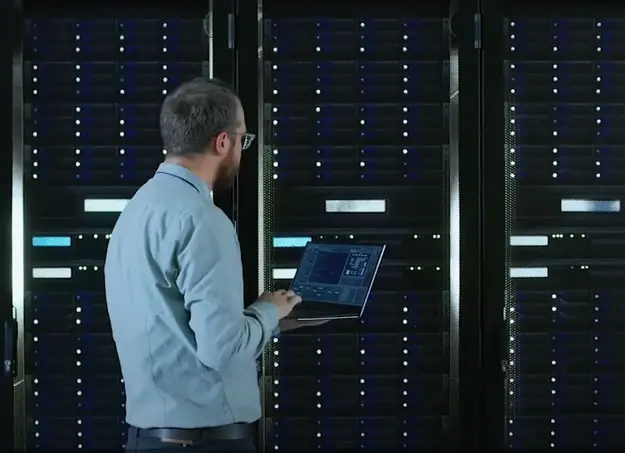 A person working in a data center or server room, using a laptop to monitor or manage the servers.