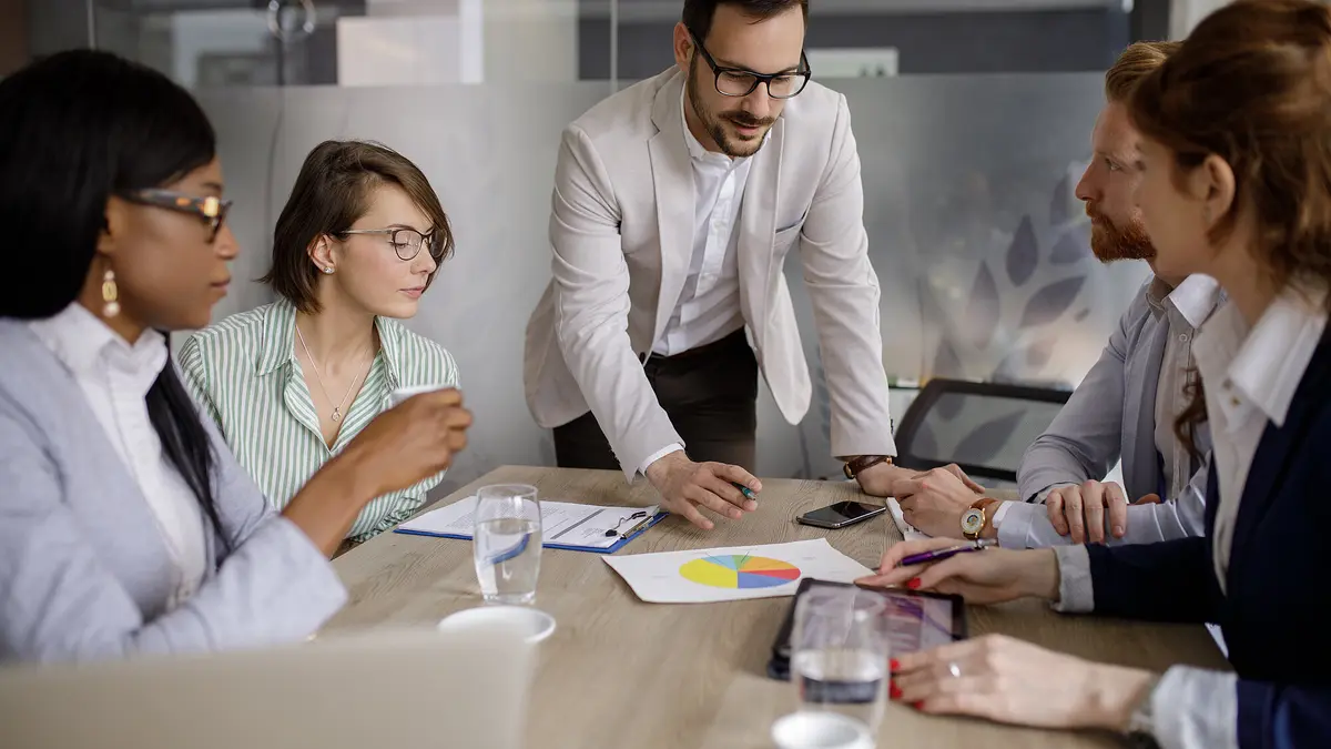 A group of professionals in a meeting, discussing around a table with laptops and documents.