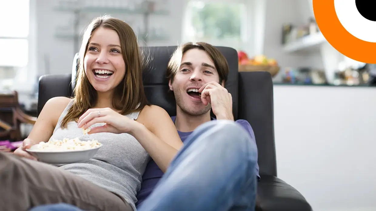Un couple assis sur un canapé consomme du pop corn en regardant un film. 