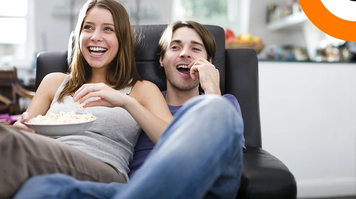 Un couple assis sur un canapé consomme du pop corn en regardant un film. 