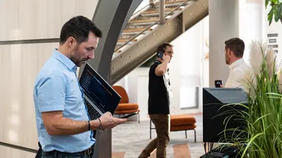 Trois hommes sont dans un bureau. Au premier plan, un homme debout tient un ordinateur sous le bras tout en utilisant son téléphone. À l’arrière-plan, un homme interpelle un autre homme qui est en communication téléphonique.