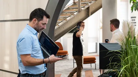 Trois hommes sont dans un bureau. Au premier plan, un homme debout tient un ordinateur sous le bras tout en utilisant son téléphone. À l’arrière-plan, un homme interpelle un autre homme qui est en communication téléphonique.
