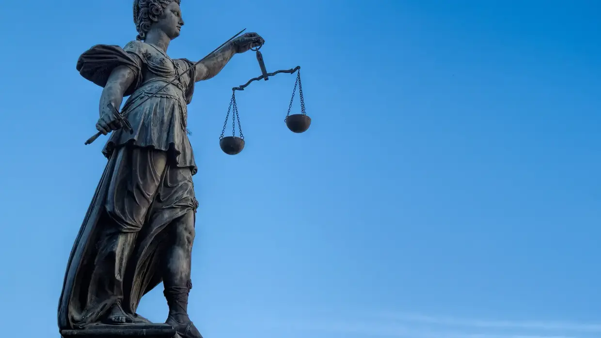 Statue einer weiblichen Figur, die eine Waage in der Hand hält, symbolisch für Gerechtigkeit. Die Statue ist aus dunklem Material gefertigt und steht gegen einen klaren blauen Himmel.