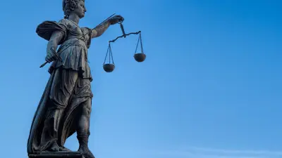 Statue einer weiblichen Figur, die eine Waage in der Hand hält, symbolisch für Gerechtigkeit. Die Statue ist aus dunklem Material gefertigt und steht gegen einen klaren blauen Himmel.