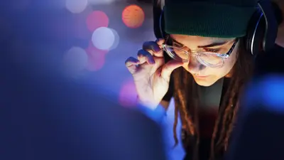 Frau mit Kopfhörern und Brille, die konzentriert auf einen Bildschirm schaut und eine Hand an die Brille hält. Der Hintergrund ist unscharf mit bunten Lichtereffekten.