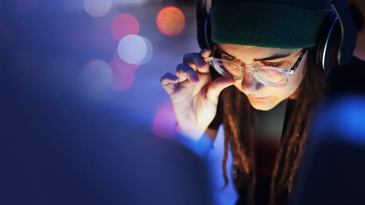 Frau mit Kopfhörern und Brille, die konzentriert auf einen Bildschirm schaut und eine Hand an die Brille hält. Der Hintergrund ist unscharf mit bunten Lichtereffekten.