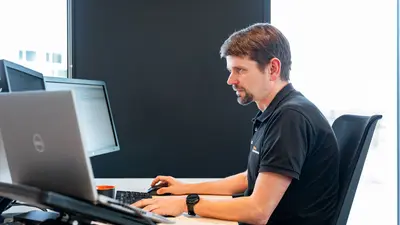 Un homme est assis à son bureau, de profil, la main droite sur la souris et la main gauche sur le clavier. Il regarde l’écran en face de lui, composé d’un double écran, avec un ordinateur portable posé à sa gauche.