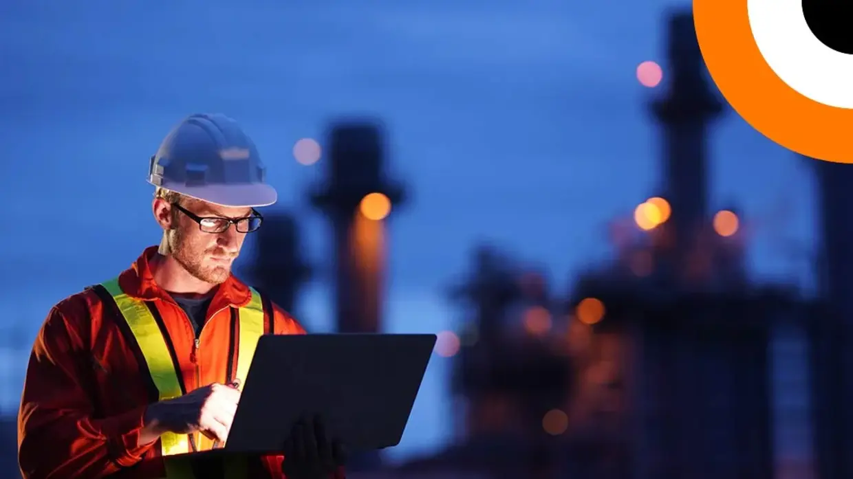Un homme vêtu d'une combinaison et d'un casque industriel consulte un ordinateur portable à la nuit tombée.