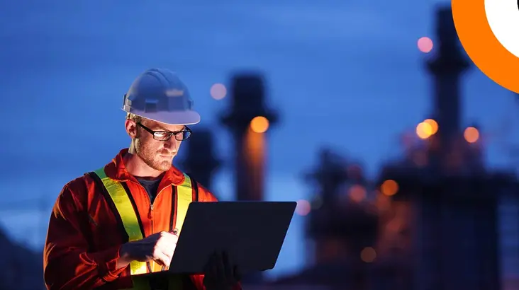 Un homme vêtu d'une combinaison et d'un casque industriel consulte un ordinateur portable à la nuit tombée.