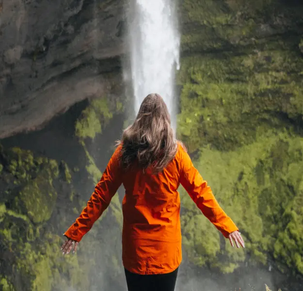 Woman in an orange jacket seen from behind with arms outstretched near a waterfall