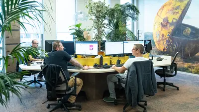 Trois hommes travaillent dans un open space organisé en cercle, chacun devant un ordinateur de bureau avec un double écran. Au centre, des plantes occupent l'espace.