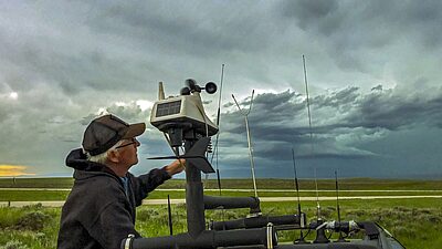 A person operating a weather or atmospheric monitoring station mounted on a vehicle, with a dramatic, stormy sky in the background.