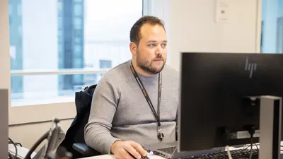 Un homme est assis de face à son bureau, une souris à la main, le regard tourné vers son ordinateur. À l’arrière-plan, on distingue une fenêtre donnant sur l’extérieur.