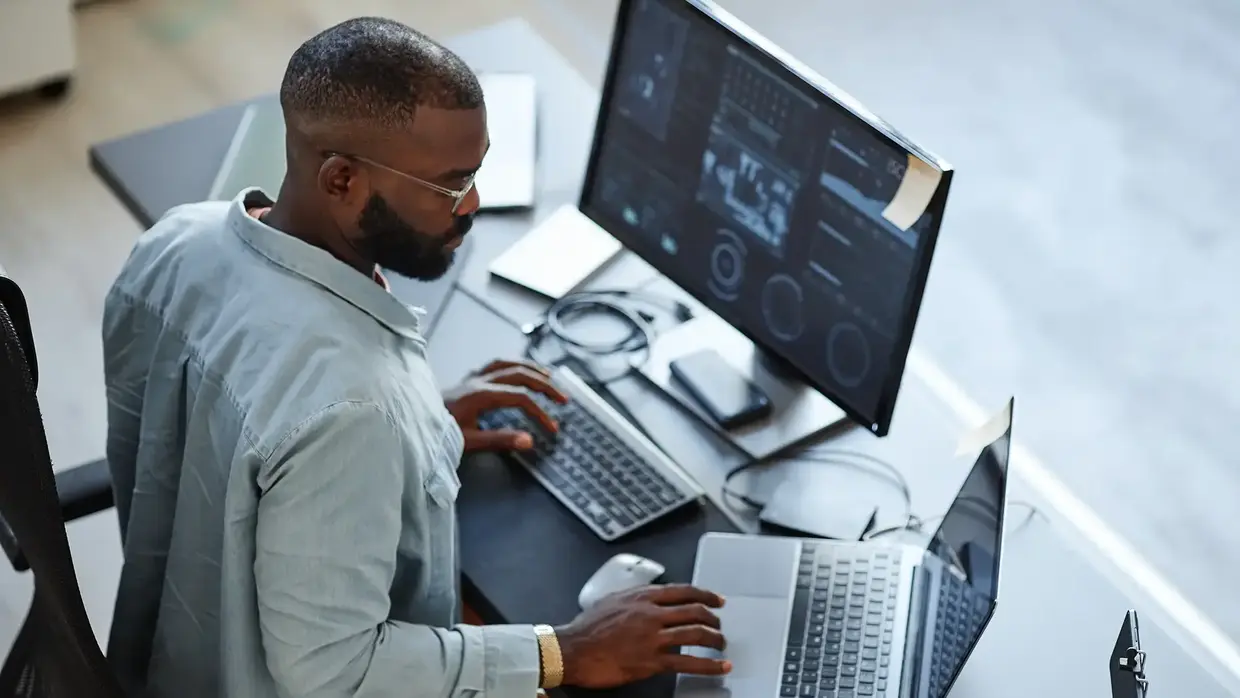 Ein Mann arbeitet an einem Schreibtisch mit mehreren Bildschirmen. Er trägt eine hellblaue Jacke, eine goldene Uhr und eine Brille. Auf dem Tisch befinden sich ein Laptop, eine Maus, ein Smartphone, Notizbücher und eine Tasse Kaffee. Der Bildschirm zeigt eine komplexe Daten- oder Analyse-Software.