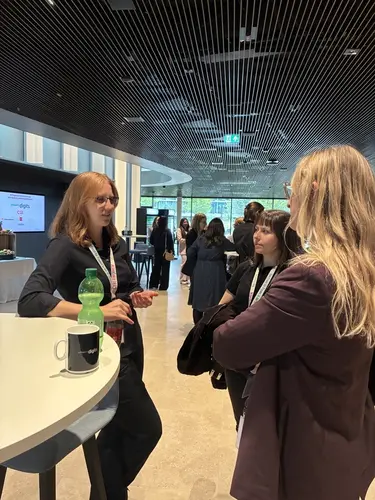 Drei Frauen stehen im Gespräch an einer Stehtheke in einem modernen, gut beleuchteten Raum mit vielen weiteren Personen im Hintergrund.