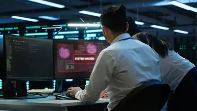 Zwei Personen in einem Serverraum oder Rechenzentrum, die an einem Arbeitsplatz mit mehreren Monitoren arbeiten. Auf einem Bildschirm ist eine Meldung zu sehen, die lautet „SYSTEM HACKED“, während der andere Bildschirm Code oder Daten anzeigt. Die Umgebung ist dunkel mit blauen und grünen Lichtern, die die Server und die technische Infrastruktur beleuchten.