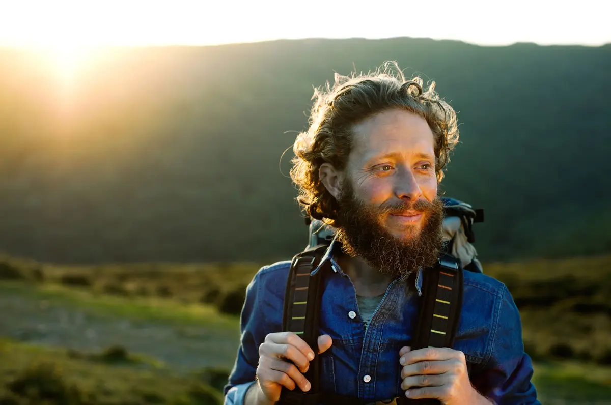 A man with a backpack, standing outdoors in a scenic landscape during sunset, smiling and looking into the distance.