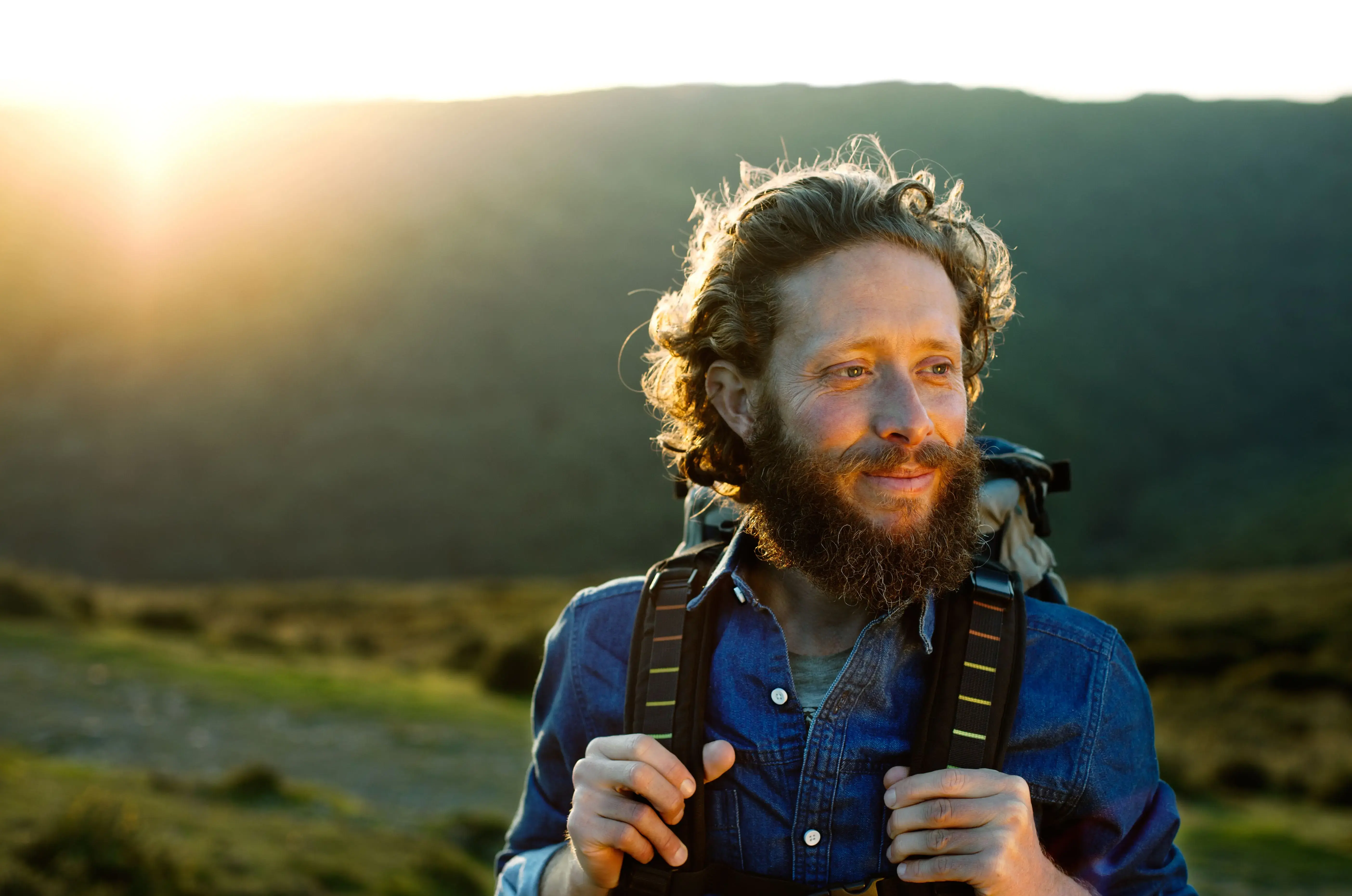 A man with a backpack, standing outdoors in a scenic landscape during sunset, smiling and looking into the distance.