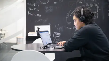 Un homme est assis dans un bureau. Il porte un casque et travaille sur un ordinateur portable. En arrière-plan, un mur noir présente des écritures à la craie.