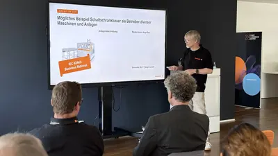 Ein Mann hält eine Präsentation vor sitzenden Personen in einem Raum mit großem Bildschirm.