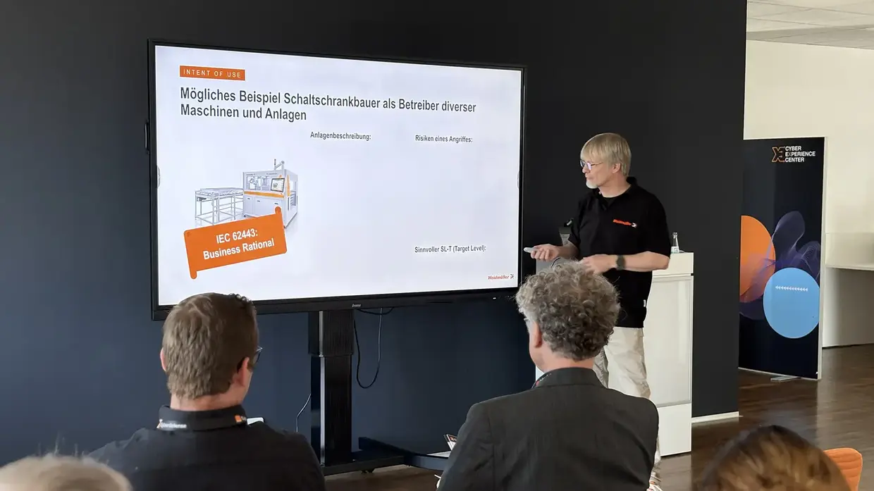Ein Mann hält eine Präsentation vor sitzenden Personen in einem Raum mit großem Bildschirm.