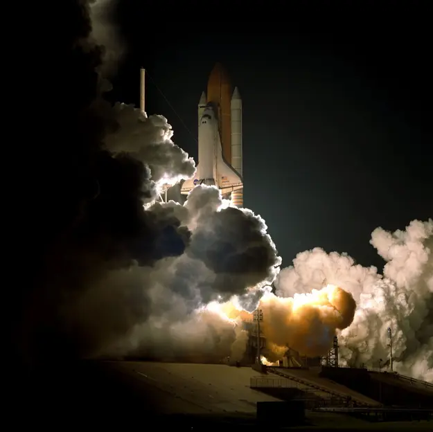 A space shuttle launching into space, surrounded by thick clouds of smoke and steam as it lifts off from the launch pad.