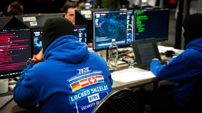 Mehrere Personen sitzen in einem Security Operations Center an Computerarbeitsplätzen mit mehreren Monitoren. Auf den Bildschirmen sind verschiedene Analyse- und Monitoring-Oberflächen sowie Code-Darstellungen zu sehen. Im Vordergrund trägt eine Person einen blauen Hoodie mit dem Aufdruck „Locked Shields 2026“.