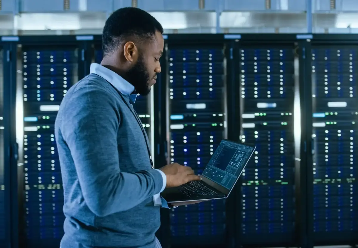 A person working in a data center or server room, using a laptop to monitor or manage the servers.