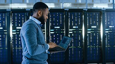 A person working in a data center or server room, using a laptop to monitor or manage the servers.