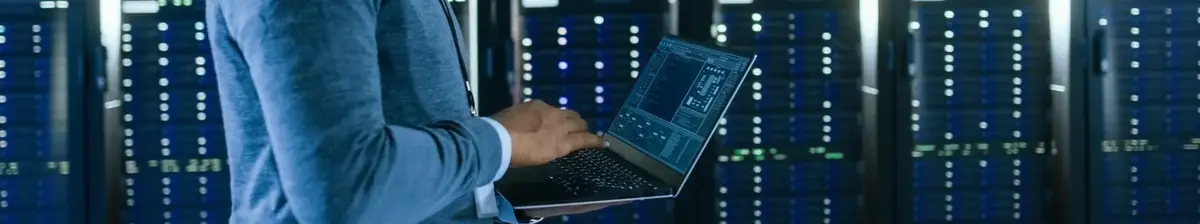 A person working in a data center or server room, using a laptop to monitor or manage the servers.