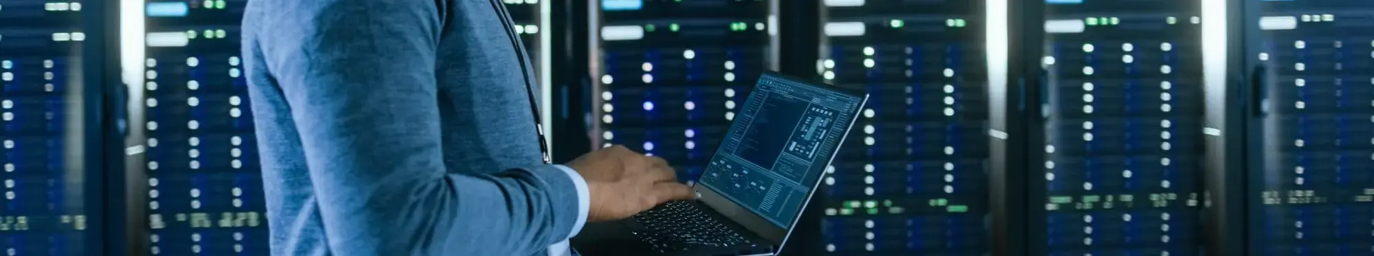 A person working in a data center or server room, using a laptop to monitor or manage the servers.