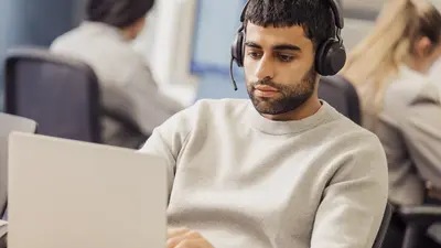 Mann mit Headset arbeitet an einem Laptop; im Hintergrund sitzen weitere Personen.
