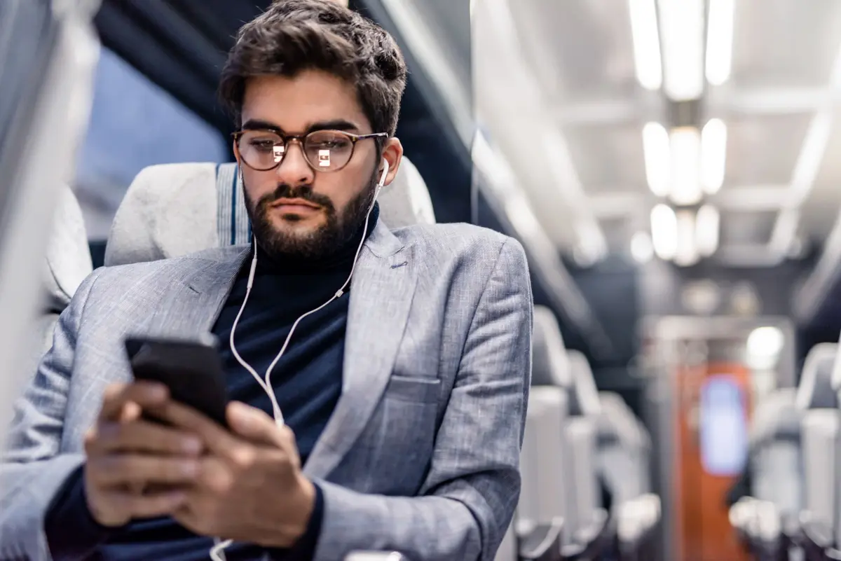 A man sitting, focused on his phone, wearing glasses and earphones, dressed in a blazer.